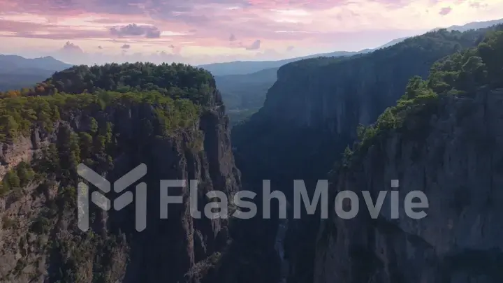 Top view of high rocks in mountainous area. Sunset sky. Pink purple clouds. The tops of the trees. Mountain river.