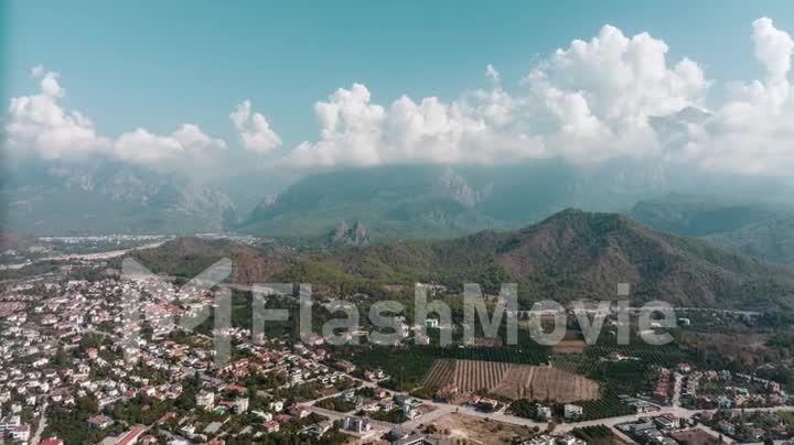 Drone footage of the city at the foot of the green mountains. Clear sunny day. White clouds. Green hills. Timelapse