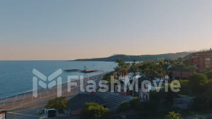 Aerial drone view of seascape at sunset. Pier. Sandy beach, umbrellas, sun loungers, palm. Mountains in the background