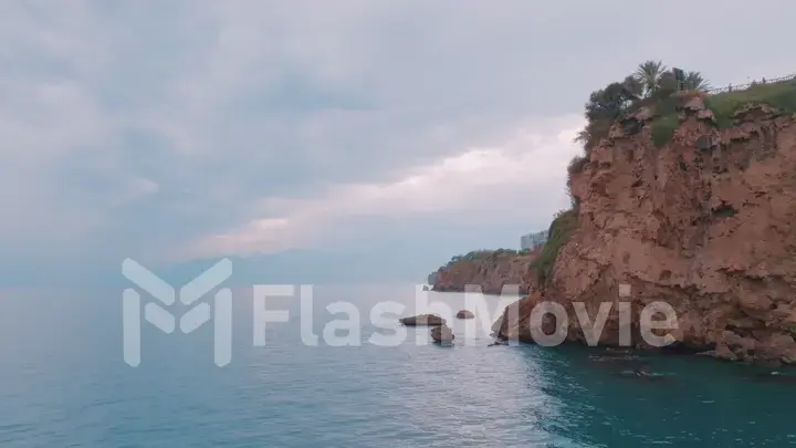 Flight over the blue sea along the shore. Red rocks. Residential buildings in the distance. Aerial drone view