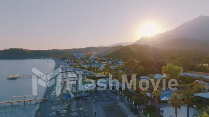 Aerial view of seascape at sunset. Pier. Sandy beach, umbrellas, sun loungers, palm. Mountains in the background