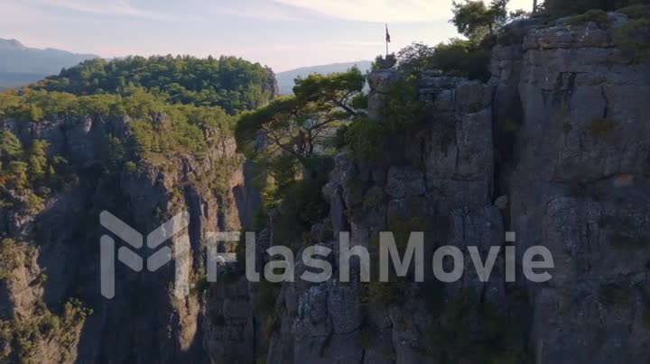 Trees grow on high gray rocks. A flag is flying on the rock. Mountain landscape. Extreme. Aerial footage from a drone