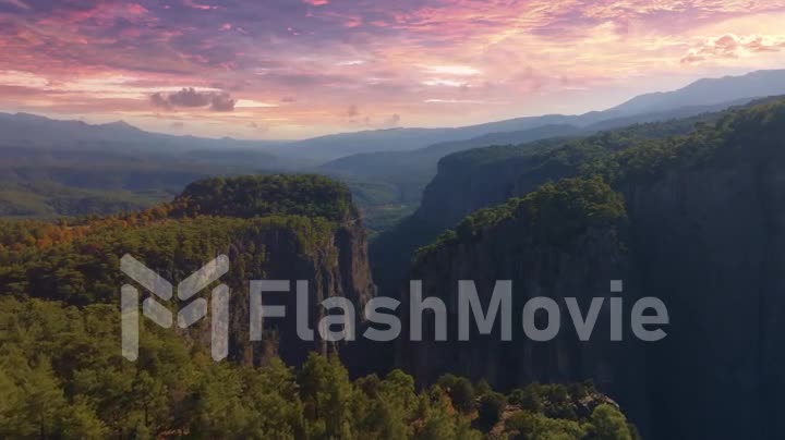 Aerial footage from a drone flying over high cliffs in a mountainous area against a sunset sky. The tops of the trees.