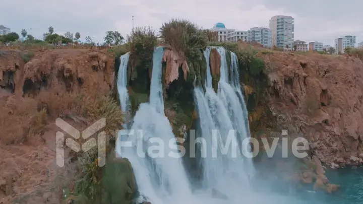 Aerial drone view of a stormy waterfall. Tall houses in the background. Red rock. Human and nature.