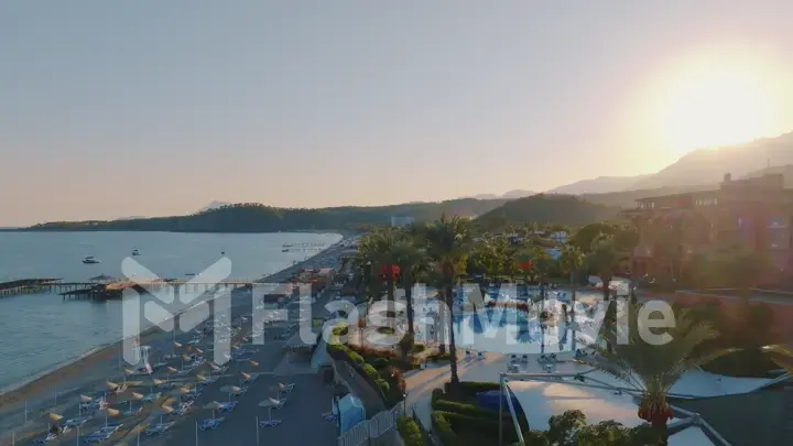 Aerial drone view of seascape at sunset. Pier. Sandy beach, umbrellas, sun loungers, palm. Mountains in the background