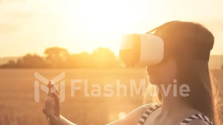 Beautiful girl in a golden wheat field uses virtual reality glasses in the sunset