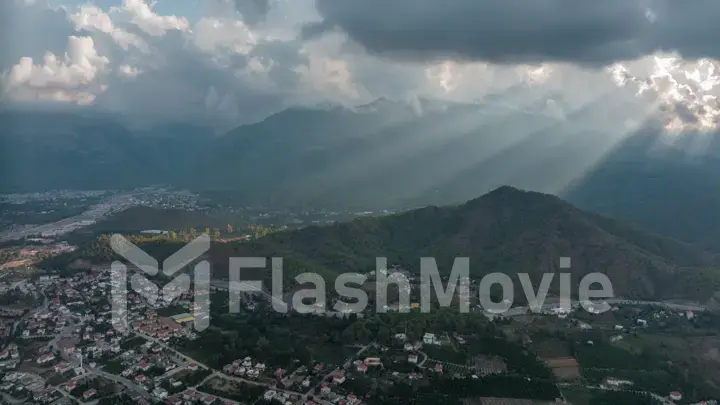 Aerial drone view of the city at the foot of the green mountains. It's a nasty day. Sunbeams between the clouds.