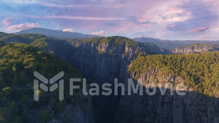 Aerial footage from a drone flying over high cliffs in a mountainous area against a sunset sky. The tops of the trees.
