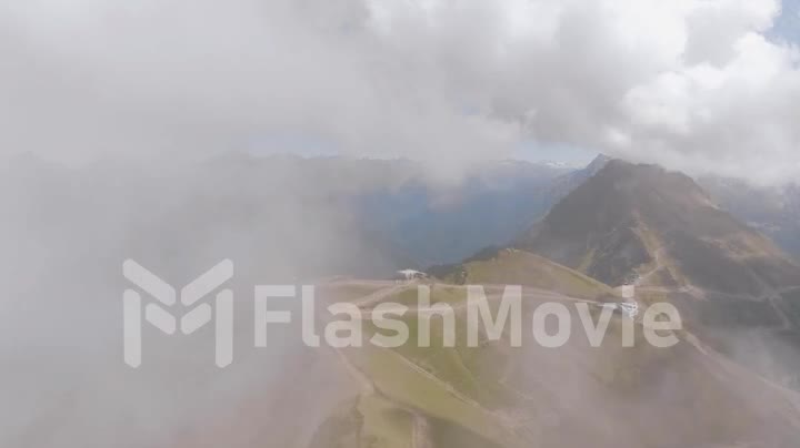 Flight through clouds over green hills on a sunny day. Roads and houses on the hill. Mountains. Drone video footage