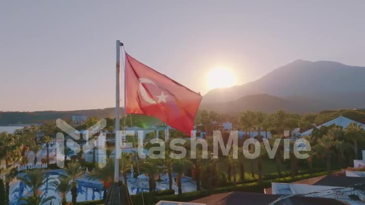 The Turkish flag is flying on the roof. Blue swimming pool, palm trees and mountains in the background. Sunset