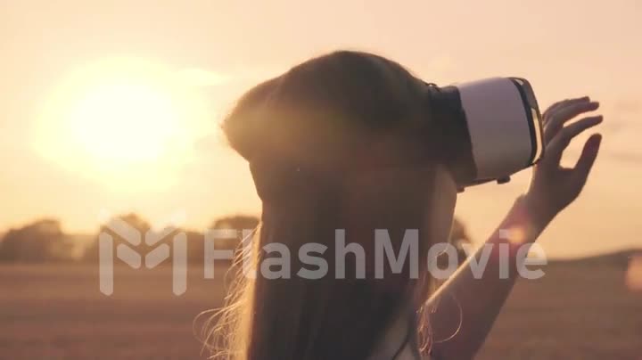 Beautiful girl in a golden wheat field uses virtual reality glasses in the sunset in slow motion