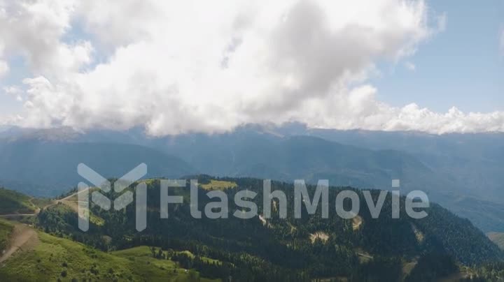 White clouds fly over the green hills. Mountain landscape. Forest on the slopes of the hills. Paths. Drone 4k footage